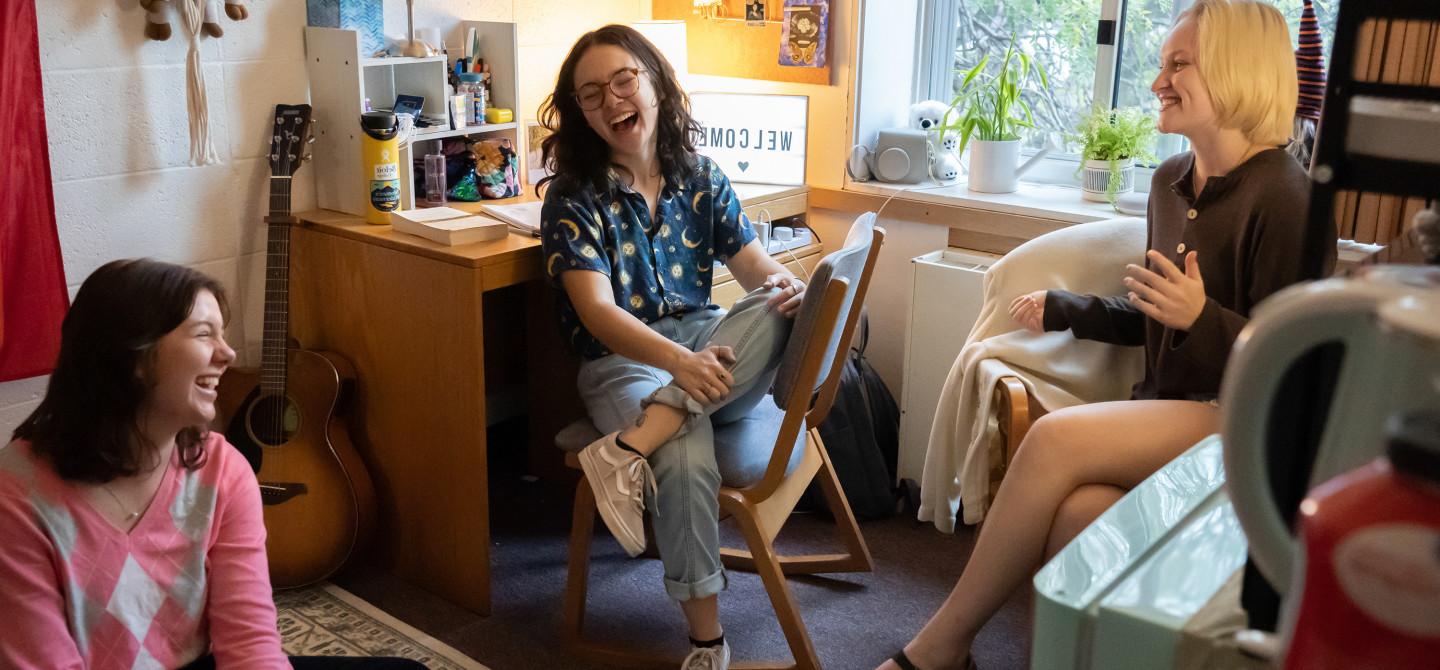 Three students hanging out together in a dorm room, laughing together.